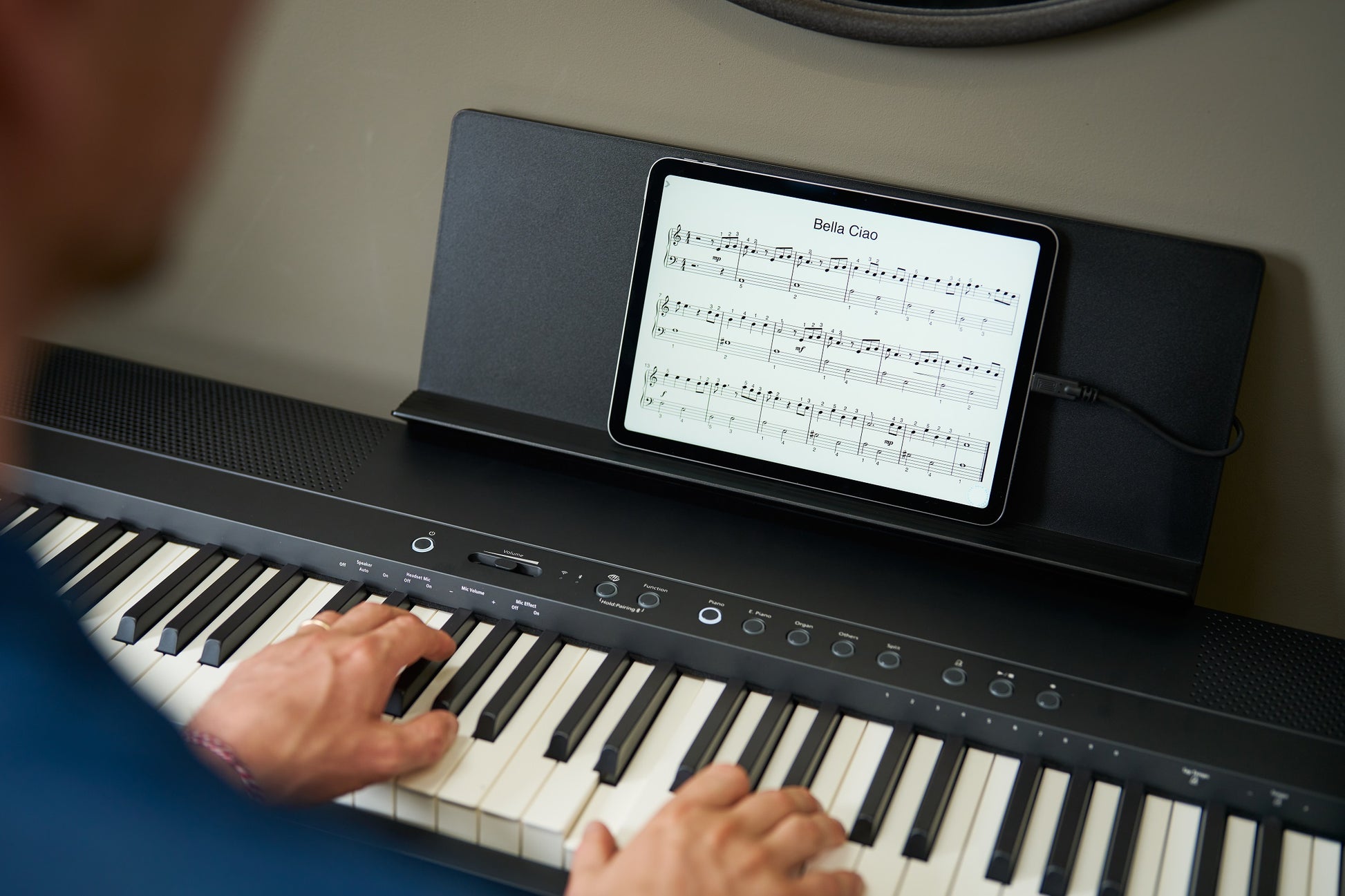 A close-up lifestyle shot of a person's hands playing a black Roland FP-40 digital piano, with a tablet displaying sheet music resting on its built-in stand against a neutral beige wall.